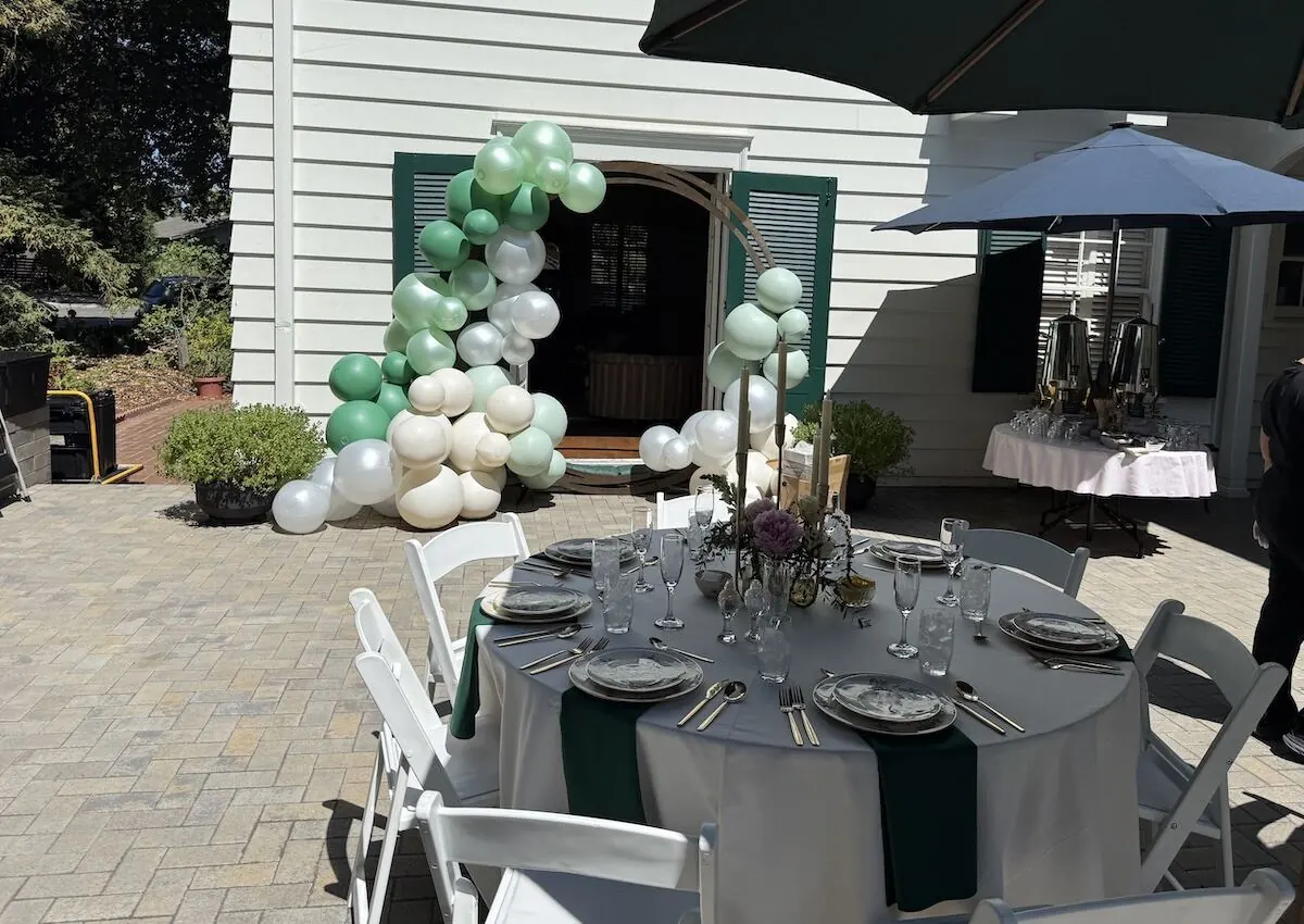 Sage and ivory balloon garland over a doorway paired with a bloom-accented tablescape — balloon and floral styling for Foster City catering parties