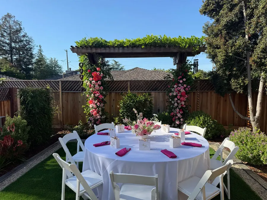 Rose arch in blush and deep red installed over a round garden table with white linen and fuchsia napkins — floral arch design for Redwood City catering weddings